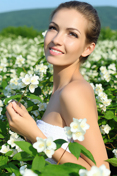 Smiling woman in white dress holding jasmine flower in blooming jasmine field under sunny sky, symbolizing the soothing and antioxidant-rich Jasmine extract in Oryskincare's rejuvenating skincare products for hydrated, youthful skin.