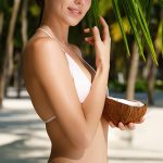 Smiling woman in white bikini holding fresh coconut under palm leaves on sunny beach, representing the natural vegetal emulsifier Montanov 68 in Oryskincare's creamy, moisturizing skincare products for stable emulsions and hydrated, smooth skin.
