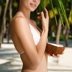 Smiling woman in white bikini holding fresh coconut under palm leaves on sunny beach, representing the natural vegetal emulsifier Montanov 68 in Oryskincare's creamy, moisturizing skincare products for stable emulsions and hydrated, smooth skin.