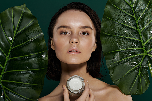 Woman holding ORY Skincare cream surrounded by lush green botanical leaves, representing botanical-infused, science-driven luxury skincare.