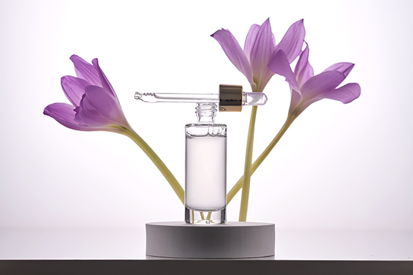 Glass serum dropper bottle containing a milky cosmetic emulsion displayed with purple botanical flowers, illustrating emulsifier science in ORY Skincare formulations.