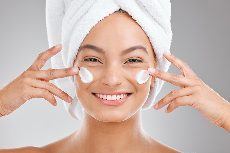 Smiling woman with towel-wrapped hair applying face cream during a refreshing ORY Skincare daily skincare routine.