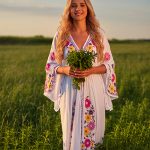Smiling woman in embroidered white dress holding fresh peppermint bouquet in sunny green field, embodying the cooling Peppermint Essential Oil in Oryskincare's invigorating skincare products for refreshed, soothed, and clear skin.