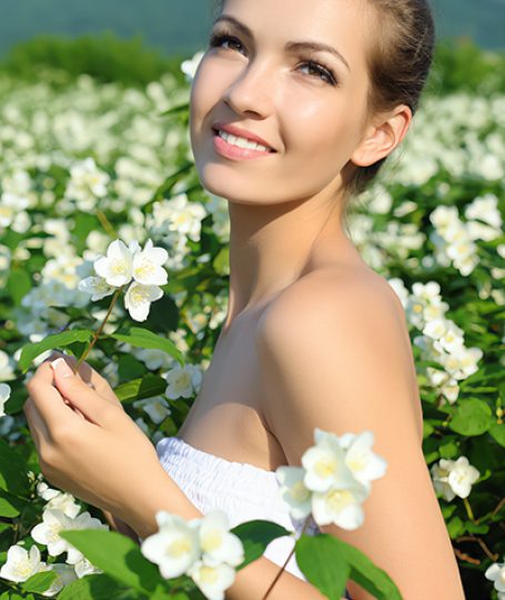 Smiling woman in white dress holding jasmine flower in blooming jasmine field under sunny sky, symbolizing the soothing and antioxidant-rich Jasmine extract in Oryskincare's rejuvenating skincare products for hydrated, youthful skin.