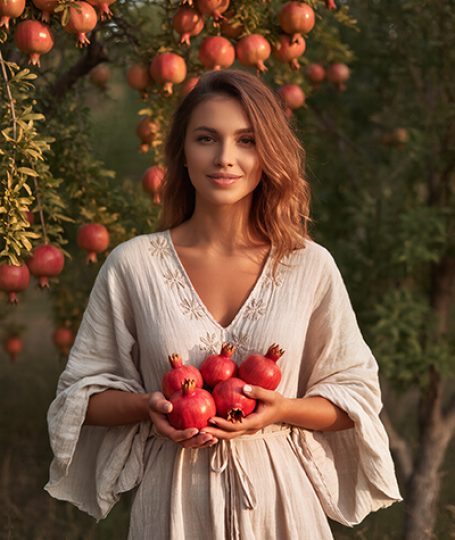 Elegant woman in beige embroidered dress holding fresh red pomegranates in lush pomegranate orchard at sunset, symbolizing the antioxidant-packed Pomegranate Extract in Oryskincare's rejuvenating skincare products for brightened, protected, and youthful skin.